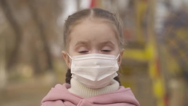 Маленькая девочка надевает медицинскую маску на лицо, защищает ребенка от коронавирусной инфекции, вспышки пандемии ковида 19, ребенок ходит по детской площадке, заботится о ребенке дыхание, детство в режиме маски — стоковое видео