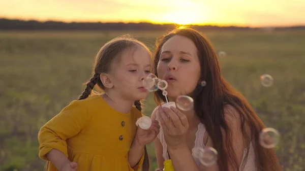 Little child with her mother blowing soap bubbles at sunset, girl daughter with mom play fun outdoors together, happy family, woman with kid laughing blowing bubbles in the air in the wind outdoors — Stock Photo, Image