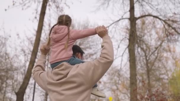 孩子坐在父亲的脖子上，爸爸背着孩子在秋天的公园里，快乐的家庭，可爱的父母，迷人的小女孩骑着露天的大自然，在天空的背景下 — 图库视频影像