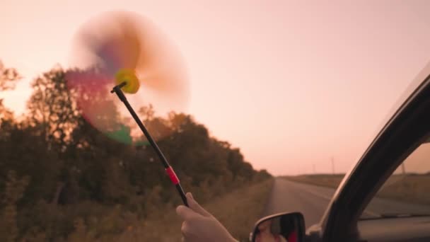 Chica libre agitando un molinete fuera de la ventana del coche en el viento, viaje camino de aventura al atardecer rosa, paseo en coche familiar, llegar por la ventana del coche a la luz del sol del cielo, disfrutar del movimiento, concepto de sueño de la infancia — Vídeos de Stock