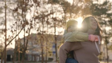Küçük çocuk annesiyle şehir parkında eğleniyor, mutlu aile hayatı, sırtında küçük bir kız, gülüyor, çocuksu arzular oynuyor, oynuyor, kızı annesine sarılıyor, gülen neşeli insanlar parlıyor.
