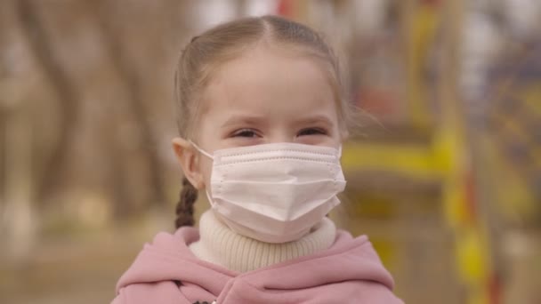 Niña se pone una máscara médica en la cara, protege al niño de la infección por coronavirus, brote de pandemia covid 19, niño camina en el patio de recreo, cuidar de la respiración del bebé, la infancia en modo máscara — Vídeos de Stock