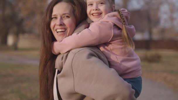 Dziecko przytula swoją matkę za szyję i śmieją się, mama trzyma małe dziecko jej ramiona i uśmiechy, szczęśliwe życie rodzinne, wesoły dziewczynka córka jazda na jej ukochanej mamie, gra z dzieckiem w parku miejskim — Wideo stockowe