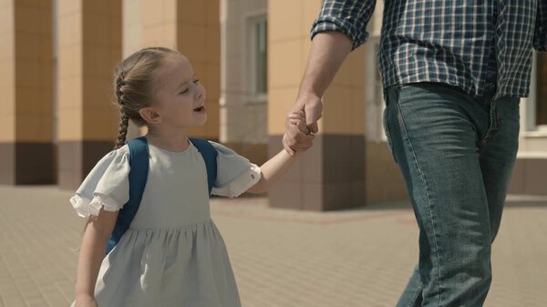 Dad is taking a little daughter to school, a girl with a backpack is holding her fathers hand, a childs school education system, a happy family, a cheerful kid with a parent walks through the