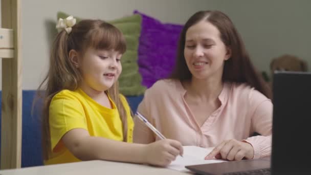 Niño pequeño escribe la tarea con un bolígrafo en un cuaderno, un niño hace la tarea con su madre en casa, aprendizaje a distancia del bebé moderno, educación preescolar con una computadora, escritorio de conocimiento del hogar de los niños — Vídeo de stock