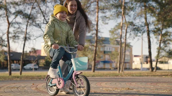 Criança aprende a andar de bicicleta de duas rodas em um parque da cidade. menina filha com sua mãe ir cavalgar na rua. vida familiar feliz. criança segura o volante enquanto dirige com pedais — Fotografia de Stock