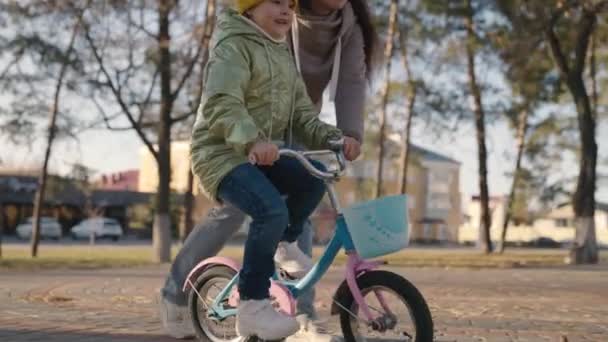 Malé dítě se učí jezdit na dvoukolovém kole v městském parku. Dcera s matkou jezdí na koni po ulici. Šťastný rodinný život. dítě drží volant při jízdě s pedály — Stock video