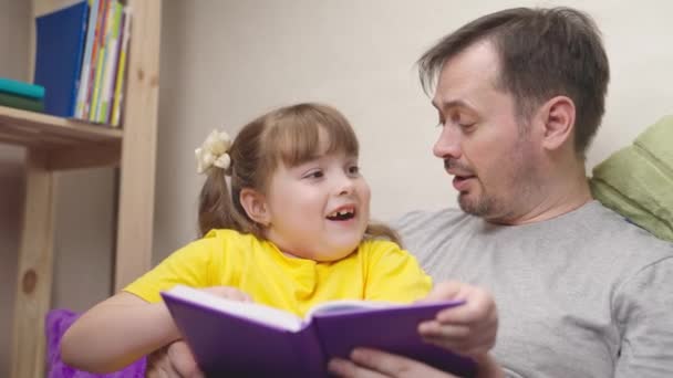 Barn läser en intressant bok med sin far, lycklig familj, flicka dotter gör läxor hemma, glad unge skrattar när du gör skollektioner, grundskoleelev lär sig att lära — Stockvideo