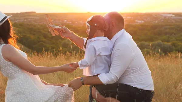 Un niño pequeño con su madre y su padre juegan un piloto de avión al atardecer, un niño en un casco sueña con volar, una familia feliz, niño de la infancia con mamá y papá en viaje, bebé hijo besa a su amada madre —  Fotos de Stock