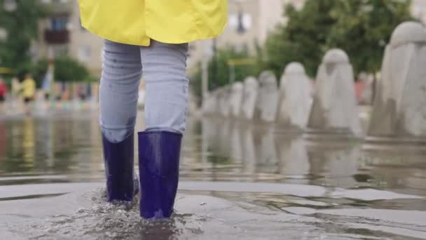 Yağmurluklu ve lastik çizmeli kızlar yağmur altında yol boyunca yürür, ayakları su birikintileri şehrinde yürür, kenarlara su sıçratır, sel caddede, araba su üstünde gider. — Stok video
