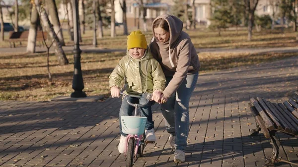 Neşeli çocuk annesiyle iki tekerlekli bisiklet sürmeyi öğreniyor, mutlu bir aile, çocuk araba sürerken pedallara gülüyor, annesiyle şehir parkında oynuyor, ebeveynler kızın ilerlemesini öğrenmesine yardım ediyor.