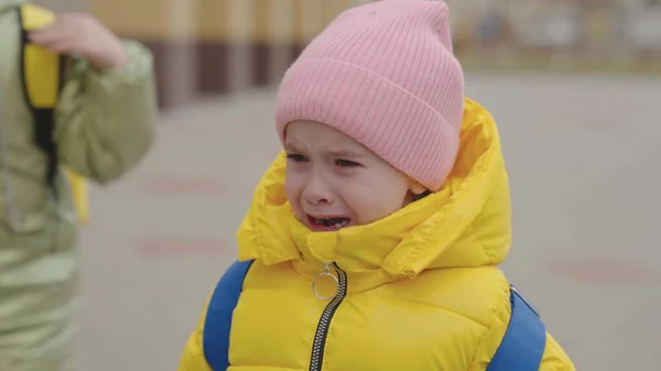 Un niño pequeño con una mochila está llorando en los brazos de su madre, las lágrimas bajan por la cara de los niños, la niña está molesta, la hija se queja con su madre, la colegiala está en la escuela con sus padres —  Fotos de Stock