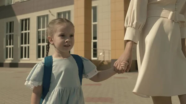 Madre lleva a un niño pequeño de la mano al primer grado de la escuela primaria, bolsa de la escuela en los hombros de una niña, familia feliz, apresurarse a la lección en el patio de la escuela, niño con mochila recibirá una educación — Foto de Stock