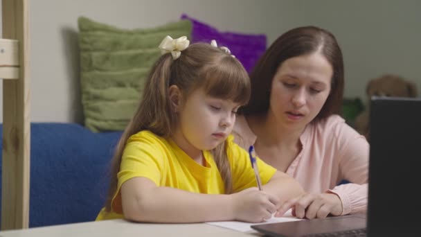 Ett litet barn gör läxor med sin mamma vid skolbänken, grabben skriver av text från bärbar skärm, mamman lär den lilla flickan att skriva i en anteckningsbok, gör fjärrlektioner hemma — Stockvideo
