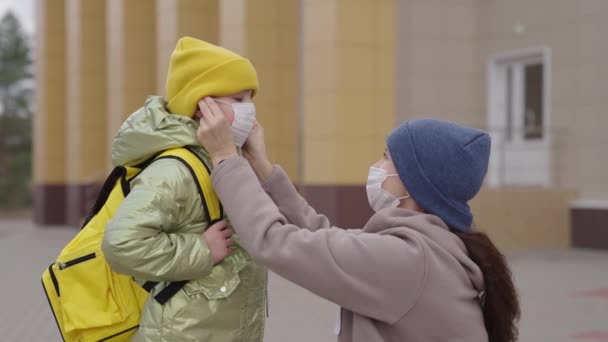 La madre se pone máscara médica en la nariz de los niños pequeños, la madre escolta al niño a la escuela, protegiendo al bebé del coronavirus durante el aula, educación en la pandemia covid-19, familia feliz — Vídeos de Stock