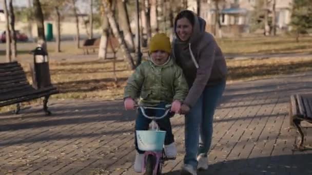 Vidám gyermek tanul lovagolni kétkerekű kerékpár az anyjával, boldog család, gyerek nevet pedál kerék vezetés közben, baby play city park az anyjával, szülő segít a lánynak tanulni előre — Stock videók