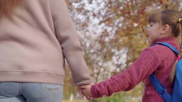 Mutter führt kleines Kind an der Hand im Herbstpark, eskortiert die kleine Tochter zur Schule, hält das Baby mit einem Rucksack in der Hand, der zum Unterricht im Klassenzimmer führt, erhält eine Ausbildung in glücklicher Kindheit — Stockvideo