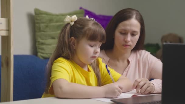 一个小孩和他的妈妈在课桌上做作业，孩子从笔记本电脑屏幕上抄写文字，妈妈教小女孩在笔记本上写字，在家上远程家教 — 图库视频影像