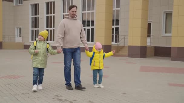 Małe dziewczynki trzymają ojca za rękę podczas spaceru z plecakami do szkoły wzdłuż ulicy miasta, dzieci otrzymują wykształcenie, szczęśliwe życie rodzinne, tata prowadzi zabawne dziecko z torbą szkolną do pierwszej klasy na lekcję — Wideo stockowe