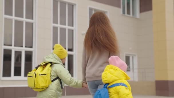 Crianças pequenas com mochilas escolares vão para a escola segurando a mão de suas mães, mamãe acompanha as filhas da criança para a aula, obter uma educação e aprender a ter sucesso na primeira série. — Vídeo de Stock
