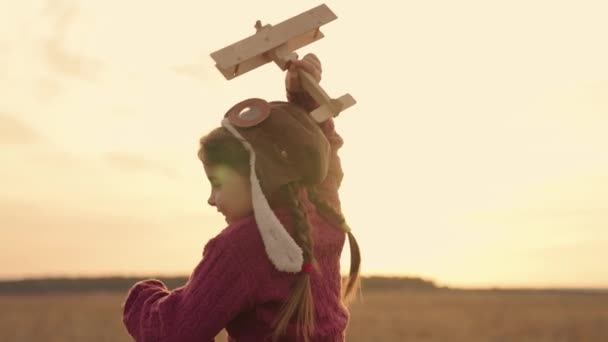 Маленький ребенок бежит с игрушечным самолетом в руке на закате, детская мечта летать, ребенок играет в пилот в шлеме, маленькие детские мероприятия на открытом воздухе, деревянные игрушки веселые девушки путешествующие лучи солнца — стоковое видео