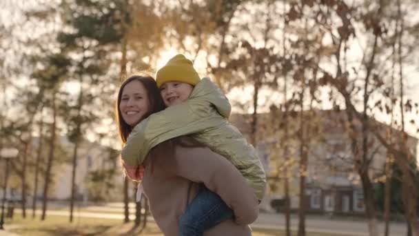 Una madre alegre rodea a su pequeña hija en sus brazos en el resplandor del atardecer de la ciudad, familia feliz, vida de maternidad, el niño con su madre se ríe abrazándose, el niño con los padres en la luz del sol, caminar parque — Vídeos de Stock