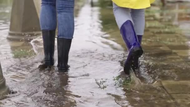 Dziewczyny w gumowych butach chodzą w kałuży, powódź na ulicy miasta, powódź po bokach dróg i asfaltowej drogi, chlapie w różnych kierunkach od mokrej błotnistej kałuży, obecność dzielnic wodnych — Wideo stockowe