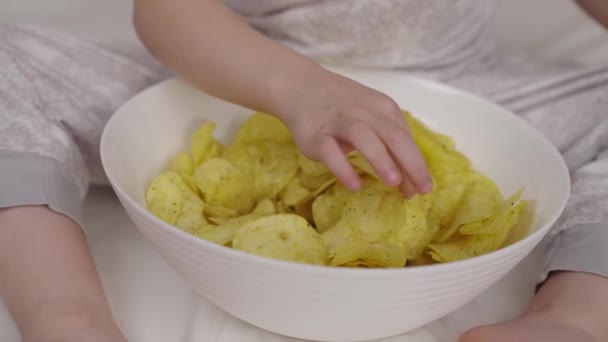 Little child eats potato chips from plate lying on bed in the bedroom, delicious food with spices and flavors for kid pleasure, crispy baby snack indoors, children food enjoying the taste of potatoes — Stock Video