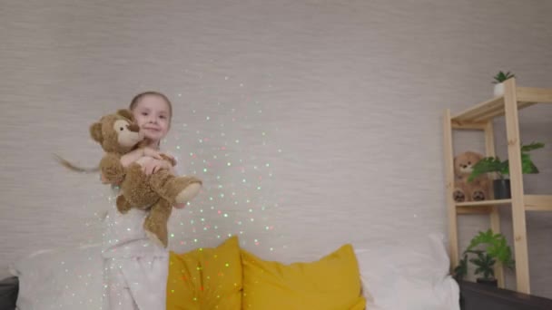 Маленька дитина стрибає з плюшевим ведмедем у ліжку в спальні, дівчинка грає з іграшкою, веселе життя танцює в яскравих різнокольорових гірляндах світла, щаслива дочка — стокове відео