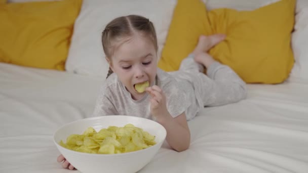 小さな子供は、ベッドの上に横たわるサクサクのフライドポテトを食べます,子供のベッドの食べ物,おいしい赤ちゃんのフードチップ,スナック,幸せな家族,家庭での子供の生活,赤ちゃんの女の子は野菜炒めを無視します,健康的ではありません人間の食べ物. — ストック動画