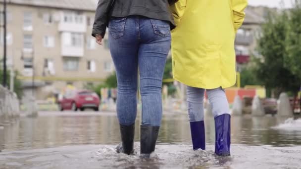 Meisjes in rubberen laarzen lopen in een plas, een overstroming op een straat, overstroming aan de zijkanten van wegen en asfaltwegen, spat verschillende richtingen uit natte modderige plas, aanwezigheid water districten — Stockvideo