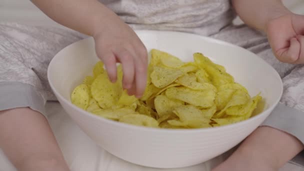 Маленький ребенок ест чипсы с тарелки, сидя на кровати, вкусный ароматный хрустящий картофель, дети быстро перекусить, вред для детей здорового питания, любовь к пряным овощным продуктам, современная семейная еда — стоковое видео