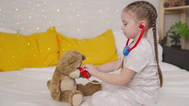 Malé dítě poslouchá medvídka se stetoskopem, dítě si hraje na doktora, zatímco sedí na posteli, dětský sen stává zdravotní sestra, veterinární lékař, dítě pomáhá zvířatům, šťastná rodina, baví hraní hraček — Stock video