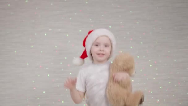 Een klein kind springt op de bank met een kerstmuts op zijn hoofd, het kind speelt met teddybeer speelgoed in het licht van felgekleurde lantaarns, bokeh bloemenslingers op kamermuur, gelukkige kinderdroom — Stockvideo