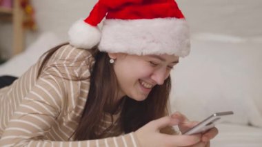 Neşeli kız sohbete ve kahkahalara bir mesaj yolluyor, mutlu noeller, Noel Baba şapkalı bir genç akıllı telefona, mutlu bir aileye bakarak gülümsüyor, gençlerin çocukluk hayalleri, yeni yıl.