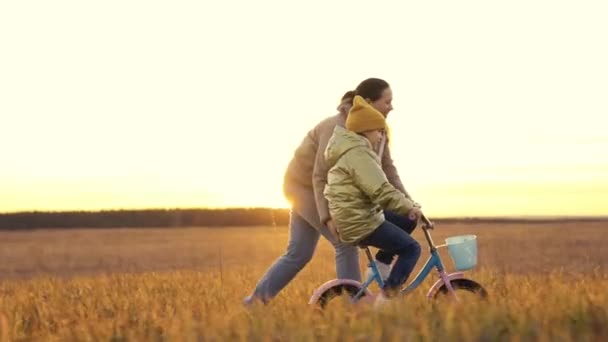 Niño aprende a montar en bicicleta con su madre, madre alegre ayuda a los niños a aprender a montar en vehículo de dos ruedas, pedales de bebé al atardecer, hija se divierte en el sol de la noche, chica con su madre esfamilia feliz — Vídeos de Stock