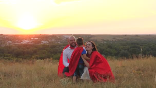 Madre y padre besan y abrazan la puesta de sol del niño pequeño, jugando a los superhéroes vestidos de capas rojas, familia feliz, valiente traje de salvador, héroes fantásticos de ensueño de la infancia, intrépido niño ganador juego divertido juego — Vídeo de stock