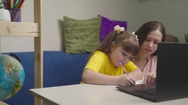 Küçük çocuk ödevlerini defterde bir kalemle yazıyor, bir çocuk evde annesiyle ödev yapıyor, modern bebek mesafesi öğrenimi, bilgisayar ile anaokulu eğitimi, çocukların ev bilgisi masası bilgisi.