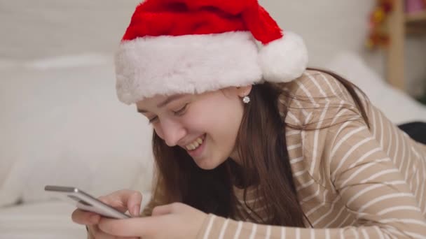 Wesoła dziewczyna wysyła wiadomość na czacie i śmieje się, Wesołych Świąt pozdrowienia, nastolatek w kapeluszu Mikołaja uśmiecha się patrząc w smartfona, szczęśliwa rodzina, nastolatki marzenie z dzieciństwa, Nowy rok — Wideo stockowe