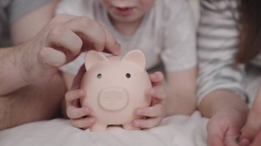 Annesi ve babası olan küçük bir çocuk domuza para fırlatıyor, mutlu bir aile kumbarasında tasarruf ediyor, çocukluk hayali konsepti, küçük bebeklerin ev parası, finansal servet tasarrufu, nakit yatırım