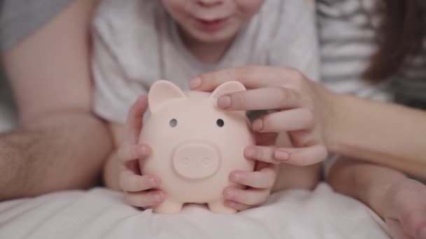 Маленька дитина з матір'ю і батьком кидає гроші на свиню, щаслива сім'я заощаджує в скарбничку, концепція дитячої мрії, маленькі немовлята гроші вдома, фінансові заощадження, інвестування готівки — стокове відео