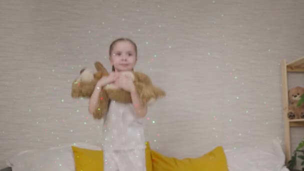 Klein kind springt op bed met teddybeer in zijn handen, gelukkig kind lacht in het licht van felle kleurrijke lichten op de muur, kleine baby speelt met zacht speelgoed, vrolijk meisje in de slaapkamer is plezier hebben — Stockvideo