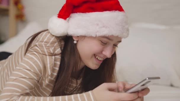 Fille gaie envoie un message au chat et rit, joyeux voeux de Noël, un adolescent dans un chapeau de Père Noël sourit en regardant dans un smartphone, une famille heureuse, un rêve d'enfance des adolescents, nouvelle année — Video