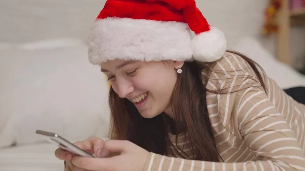Munter pige sender en besked til chatten og griner, glædelig jul hilsner, en teenager i en santa hat smiler ser ind i en smartphone, en lykkelig familie, en teenagere barndomsdrøm, nytår Stock-foto