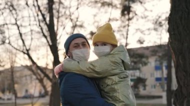 Maske takmış küçük bir kız, babasının kollarında tıbbi maskeli bir kız güneşli bir şehirde gülümsüyor, gün batımında babasıyla birlikte, kendisini koronavirüs salgınından korumak için, covid 19, kod gülüyor.