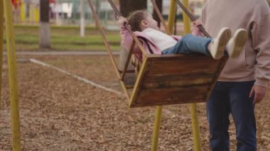 Anne küçük çocuğu sallıyor salıncakta, neşeli çocuk yukarı aşağı uçuyor, bebek oyun oynarken gülüyor ve gülümsüyor, anne kız parkta yürüyor, mutlu aile, çocukluk hayali uçuyor.