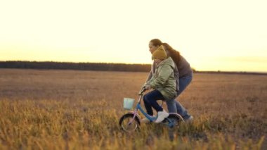 Küçük çocuk annesiyle bisiklet sürmeyi öğreniyor, neşeli anne çocuğa iki tekerlekli araba sürmeyi öğretiyor, gün batımında bebek pedalları, akşam güneşinde kız ve annesi mutlu bir aile.