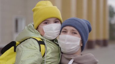 Annesi sırt çantalı ve yüzünde sağlık maskesi olan küçük okul kızına sarılıyor, 2019 Covid virüsü, okulda eğitim alırken, anne çocuğa ders için birinci sınıfa kadar eşlik ediyor, koronavirüs salgını