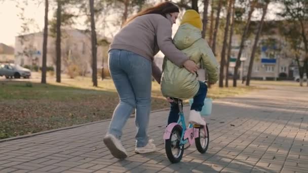 Matka učí malé dítě jezdit na kole ve městě, dítě jezdí na chodníku, šťastná rodina, dítě hraje a směje se s mámou, učit se jezdit, aby se dětský sen stal skutečností dívky, dcera a máma jsou zábavné — Stock video