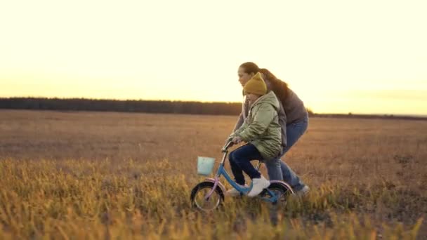 Klein kind leert fietsen met zijn moeder, vrolijke moeder helpt kind leren rijden tweewielige voertuig, baby pedalen bij zonsondergang, dochter heeft plezier in de avondzon, meisje met haar moeder ishappy familie — Stockvideo